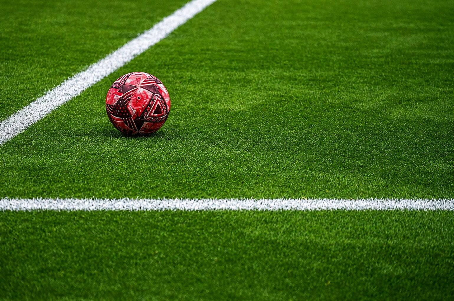 Ballon de football rouge posé sur une pelouse verte avec lignes blanches visibles sur un terrain de sport