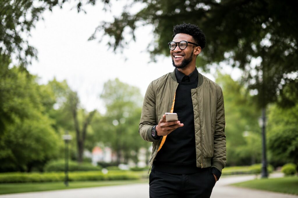 Jeune homme souriant tenant un smartphone lors d'une promenade dans un parc verdoyant.